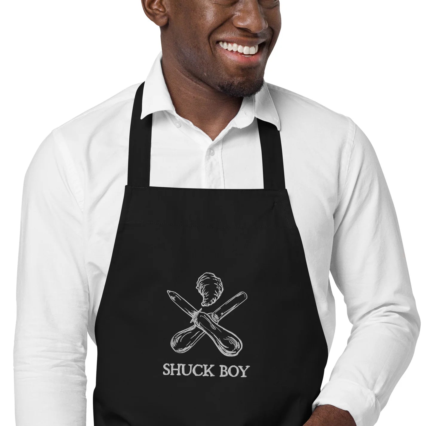 Cropped view of man in a white button-down shirt with an organic apron on. The apron has "SHUCK BOY" and an oyster knife and a clam knife crossed in an "x" design with an oyster on top done in embroidery. Oyster clothing for shuck boys, shuck babes.