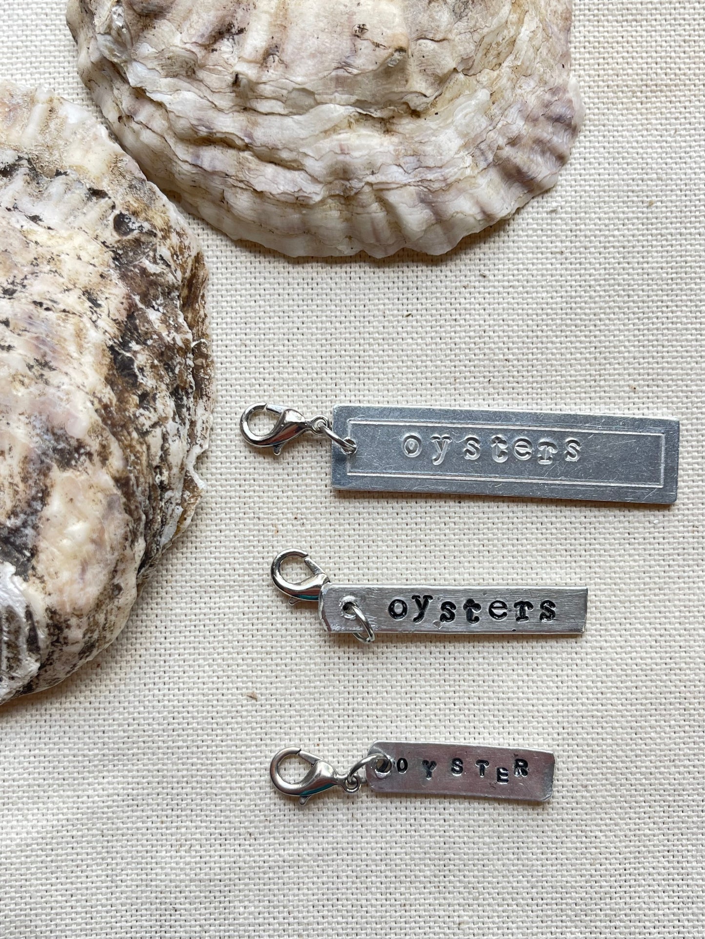 Three silver 'oyster' charms on a textured surface with oyster shells in the background.